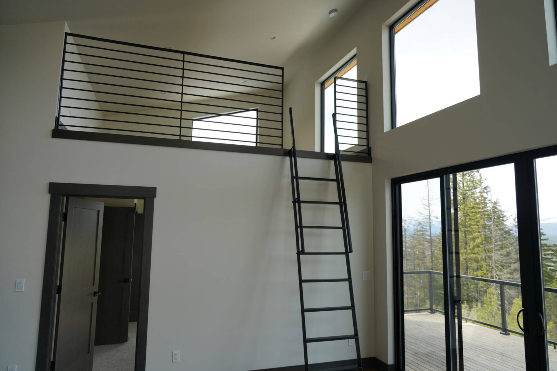 A modern room with a black metal ladder leading to a loft area with metal railings, large windows letting in natural light, and a glass door opening to a balcony overlooking trees.