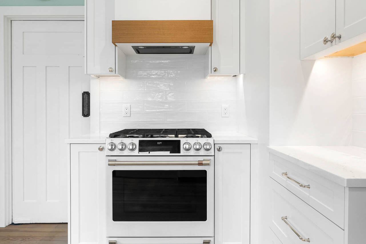 Modern white kitchen with a stainless steel gas oven and stovetop, wood-accented range hood, white cabinets, light wood floors, and a wall clock above a closed door.