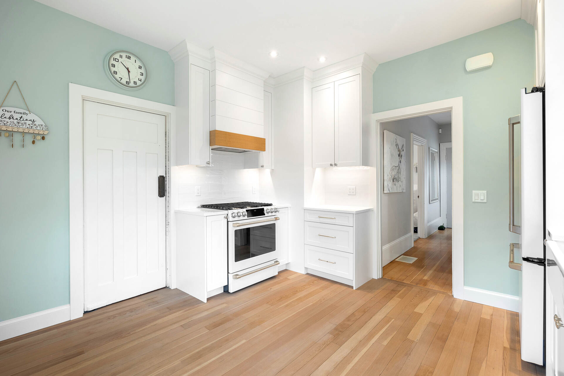 Bright kitchen with light wood floors, white cabinets, a white stove, a door with a black handle, a wall clock, and light blue walls. A hallway with wood flooring is visible in the background.