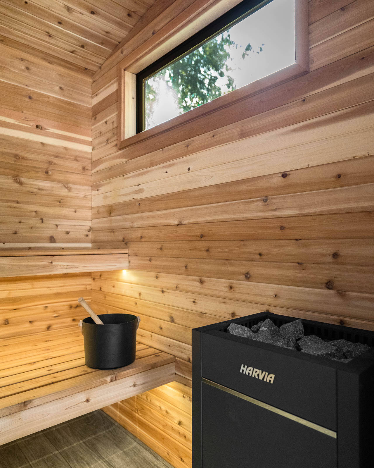 A modern wooden sauna interior with a bench, a black bucket and ladle, a Harvia heater with stones, wood-paneled walls, and a rectangular window showing trees outside.