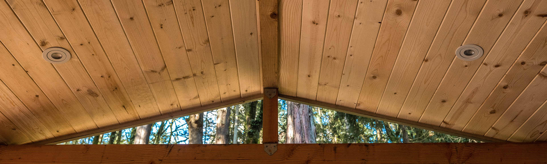 View of a wooden, vaulted ceiling with exposed beams, seen from below. Behind the ceiling, tall trees and green foliage are visible through an open space.