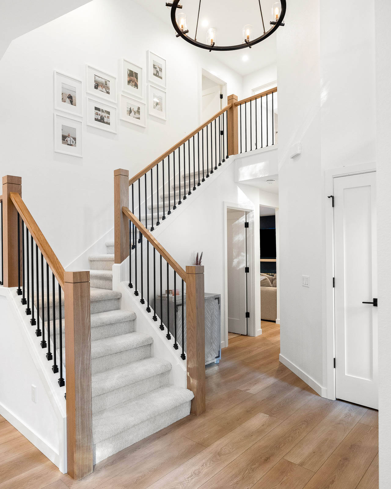 A modern, bright foyer with a wooden staircase featuring black railings, light wood floors, white walls, a round chandelier, and framed photos on the wall above the stairs.