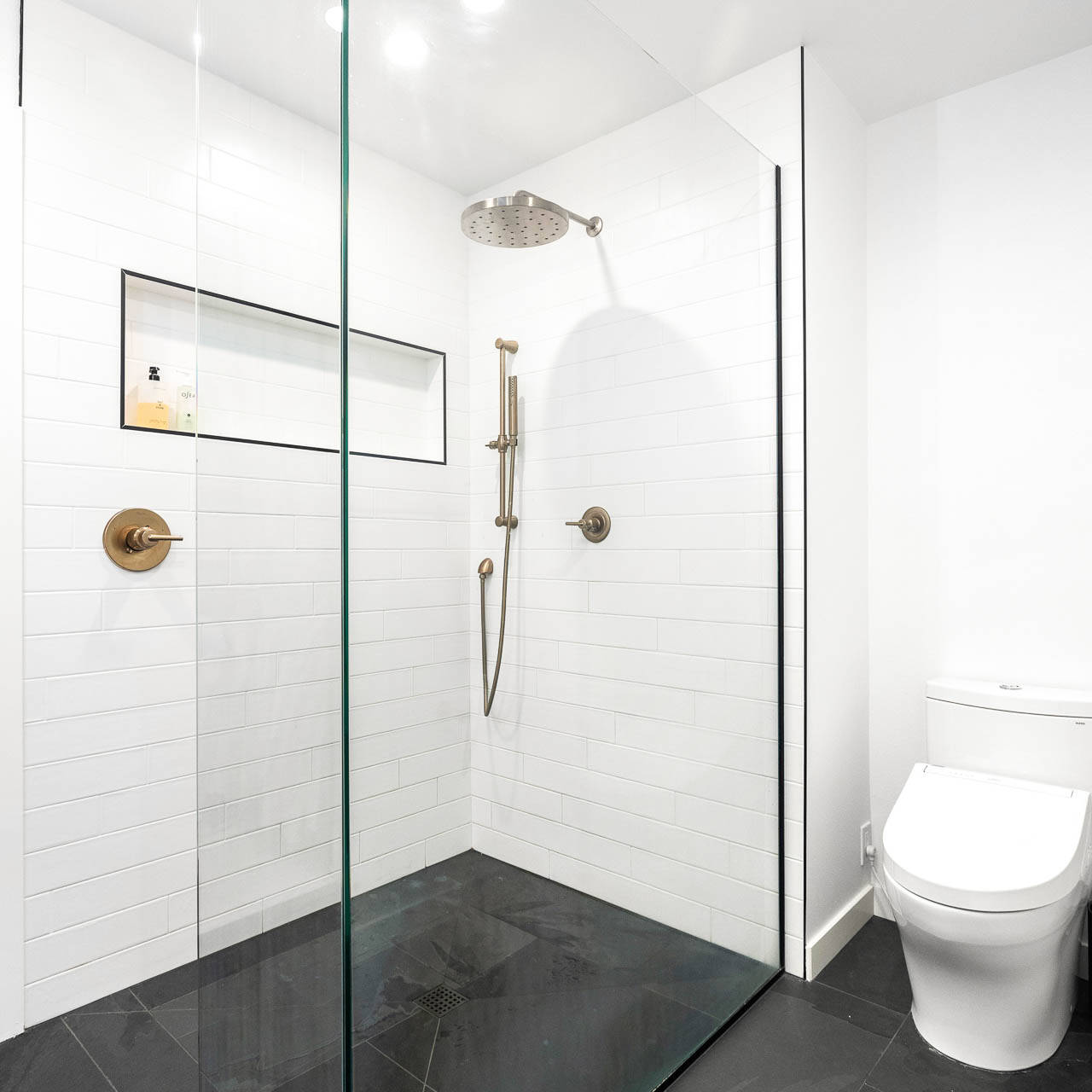 A modern bathroom with a frameless glass shower enclosure, white subway tile walls, a rainfall showerhead, black floor tiles, and a white toilet beside the shower.