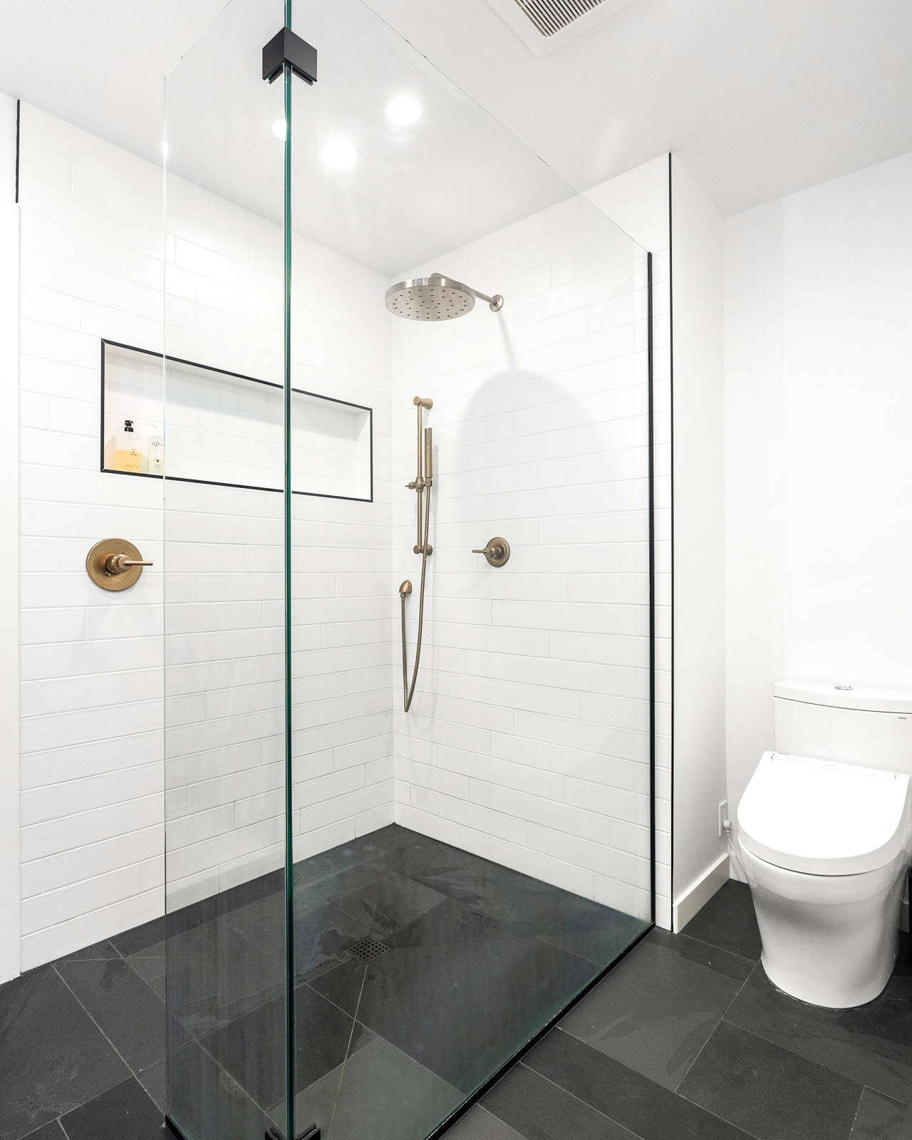 A modern bathroom with a frameless glass shower enclosure, white subway tile walls, a rainfall showerhead, black floor tiles, and a white toilet beside the shower.