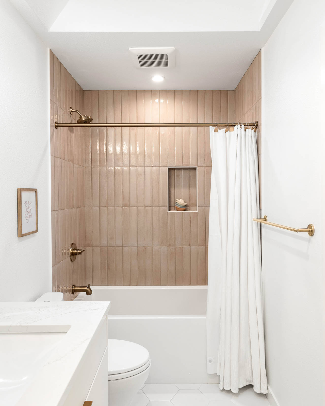 A bright, modern bathroom features light pink vertical tiles around a bathtub with gold fixtures, a white shower curtain, white walls, hexagonal floor tiles, and a small framed picture on the wall.
