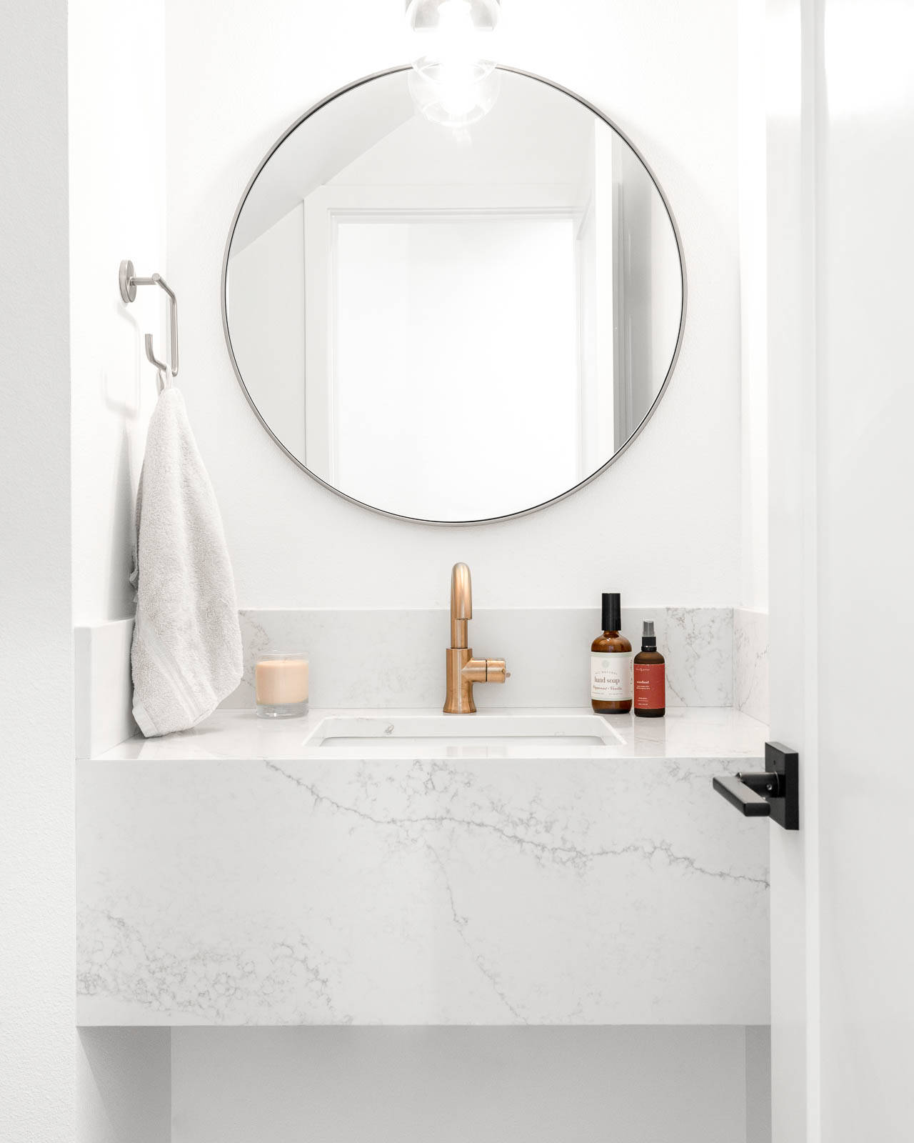 A modern bathroom with a white marble sink, gold faucet, round mirror, a hanging towel, and bottles of soap and lotion on the counter. A lit candle and overhead light add a cozy touch.