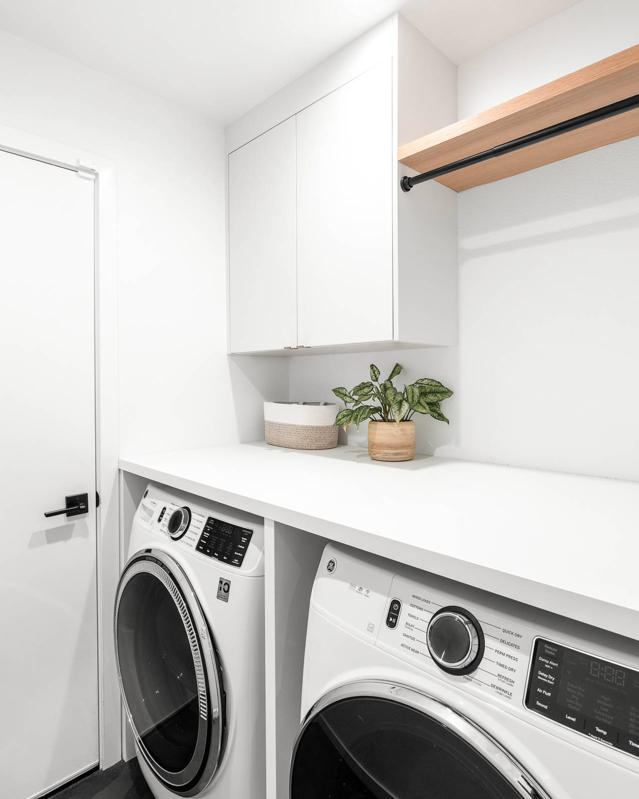 A modern laundry room with a white washer and dryer side by side, white cabinets above, a wooden shelf with a black rod, and two potted plants on the counter. The room has white walls and a black floor.