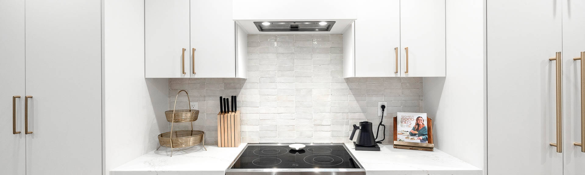 Modern kitchen with white cabinets, an electric stove, a vent hood, a knife block, a kettle, stacked baskets, and a cookbook on the countertop. The backsplash features light-colored tiles.