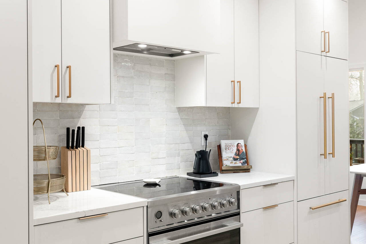 Modern kitchen with white cabinets, gold handles, a stainless steel oven, white countertops, backsplash tiles, a knife block, and a kettle. Wooden floors and natural light from a window enhance the bright space.