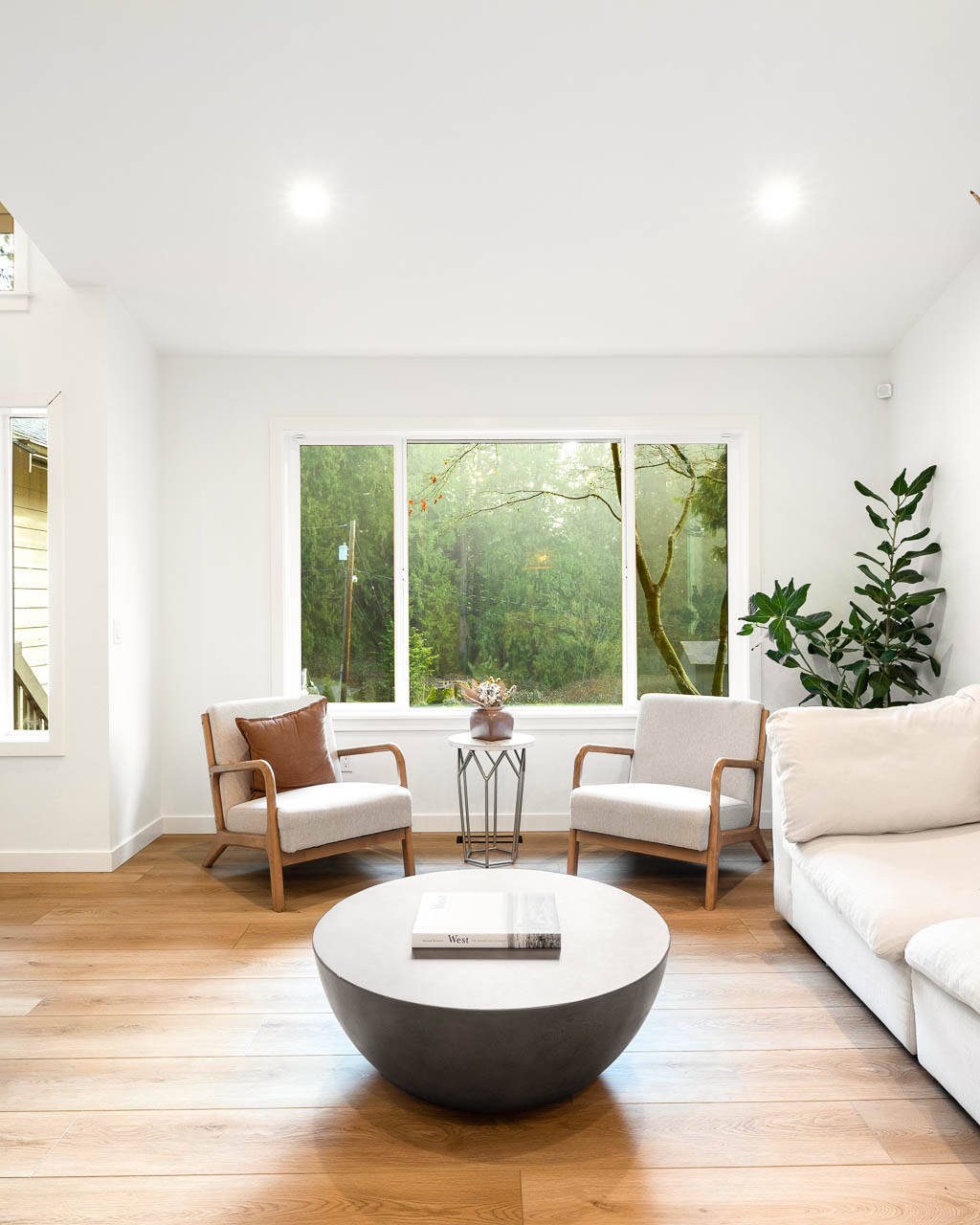 A modern, bright living room with light wood floors, a white sofa, two beige chairs, a round coffee table, large window, indoor plants, and a wall-mounted deer antler decor.