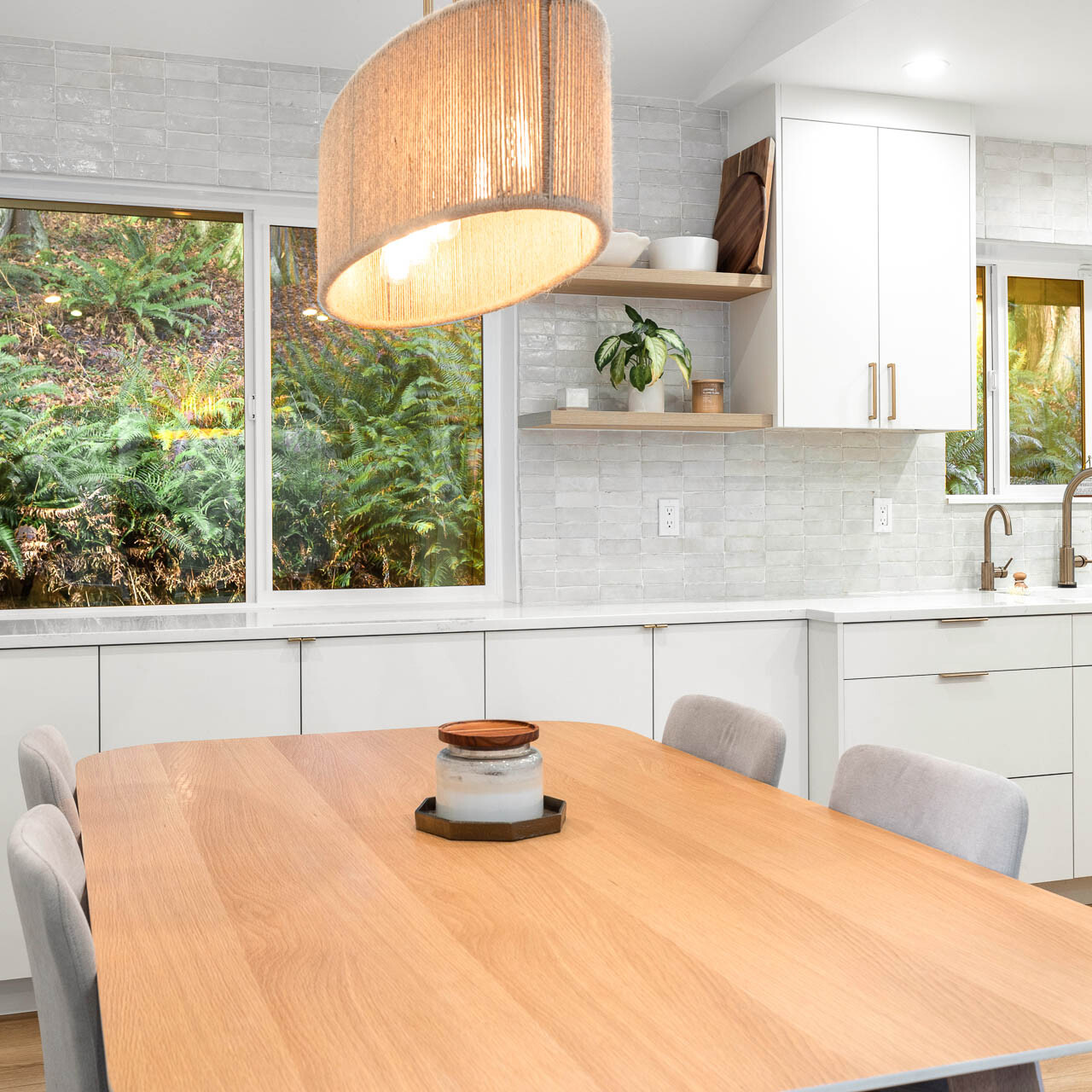 A modern kitchen with white cabinets, a light wood dining table, cushioned chairs, open shelves, a large pendant lamp, and windows showing greenery outside. The space is bright and tidy with minimalist decor.