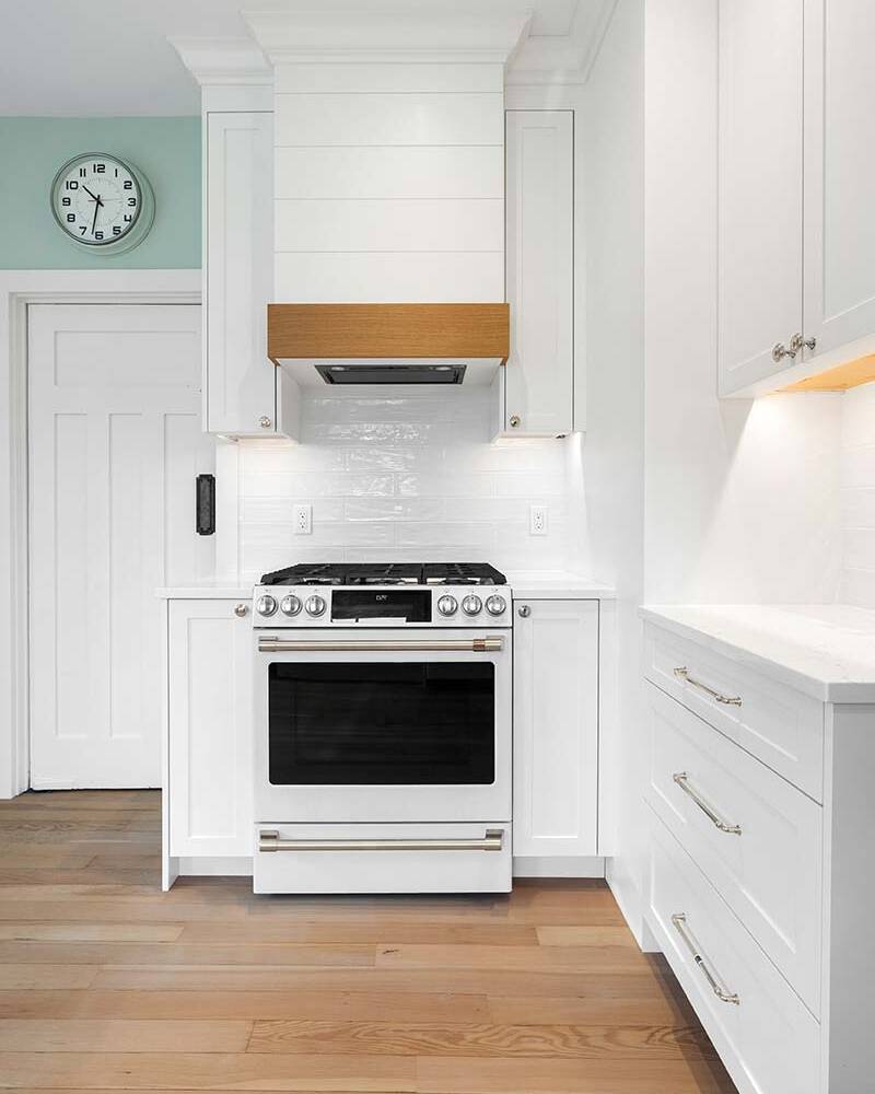 A modern kitchen features a white gas stove with a wooden range hood, surrounded by white cabinets and drawers. Light wood flooring and a wall clock complete the clean, minimal design.