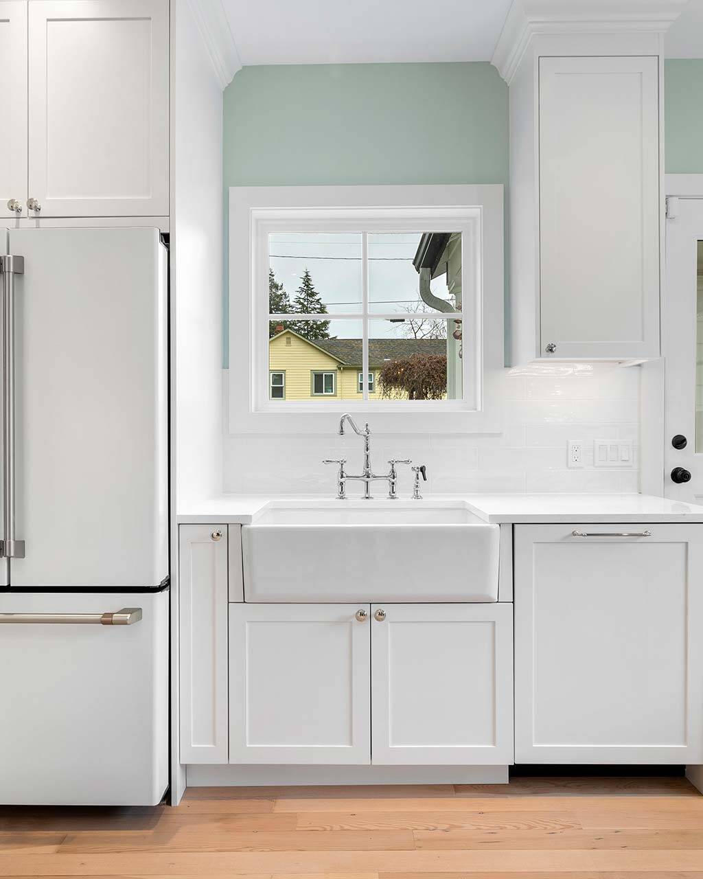 A bright, modern kitchen with white cabinets, a farmhouse sink under a window, a white refrigerator, light wood flooring, and a white door leading outside. Houses and trees are visible through the window.