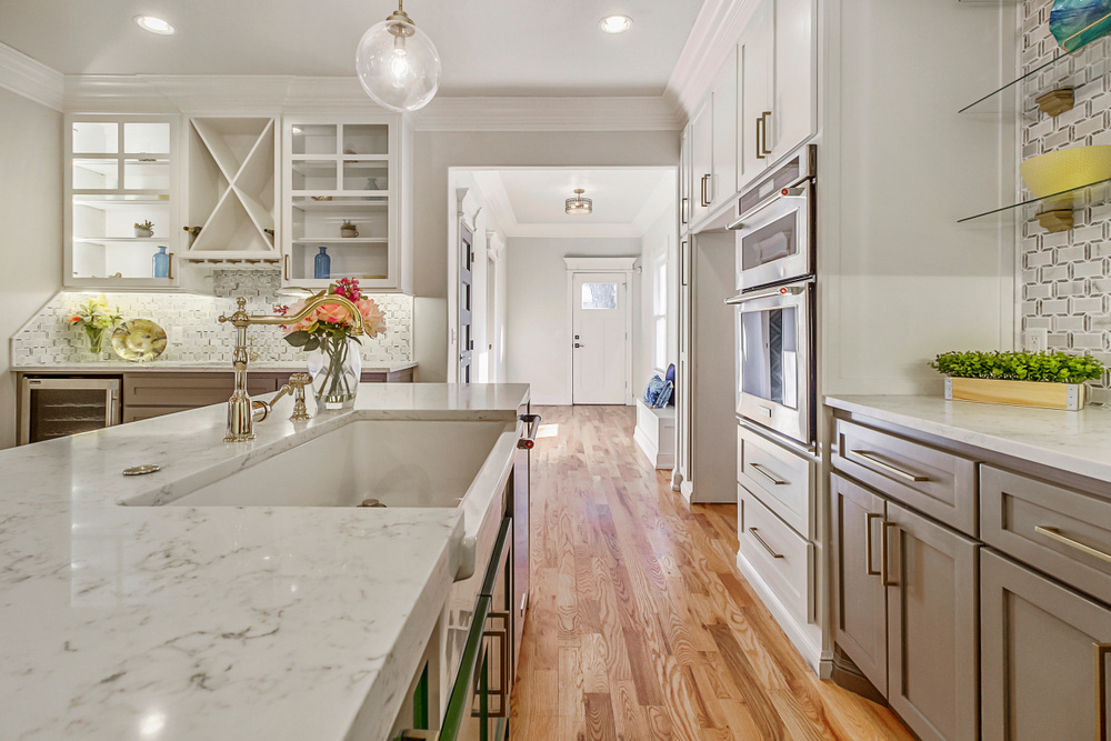 Modern kitchen with white marble countertops, light wood floors, gray cabinets, built-in ovens, glass-front cabinets, and decorative flowers on the island under bright lighting.