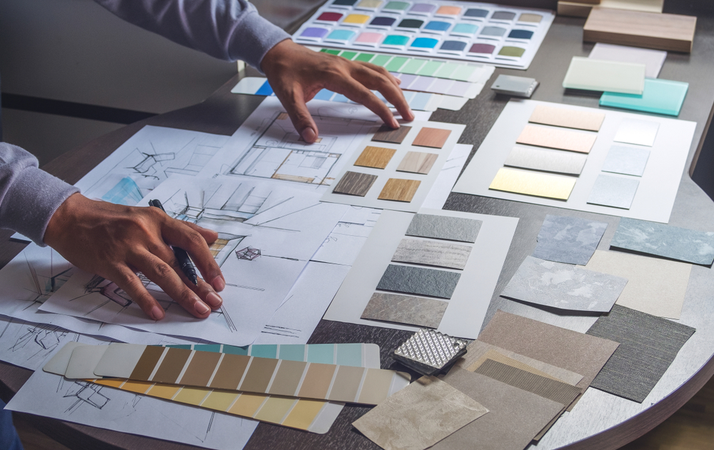 Close-up of a person’s hands working on interior design plans, surrounded by color swatches, material samples, and architectural drawings spread out on a table.