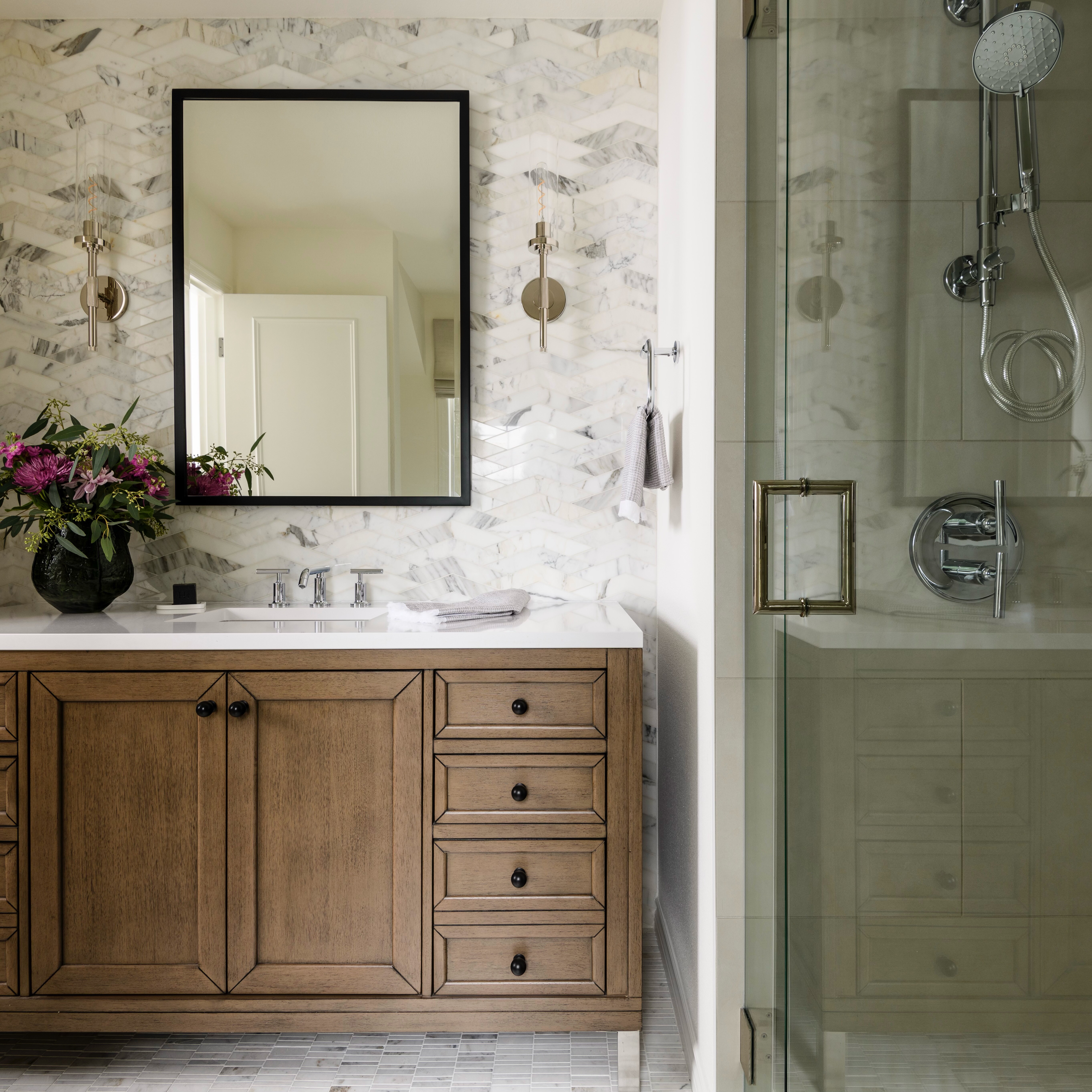 Modern bathroom with a wooden vanity, marble herringbone backsplash, rectangular mirror, and glass shower. A vase with flowers sits on the counter and two towels hang on the wall.