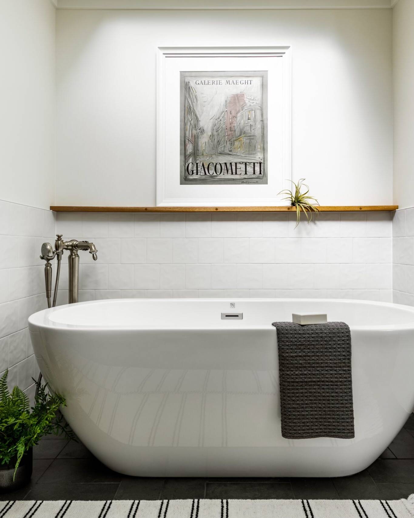 A modern white bathroom with a freestanding bathtub, a gray towel draped over the tub, a potted fern on the floor, a striped rug, wall art above the tub, and a white towel hanging on the wall.