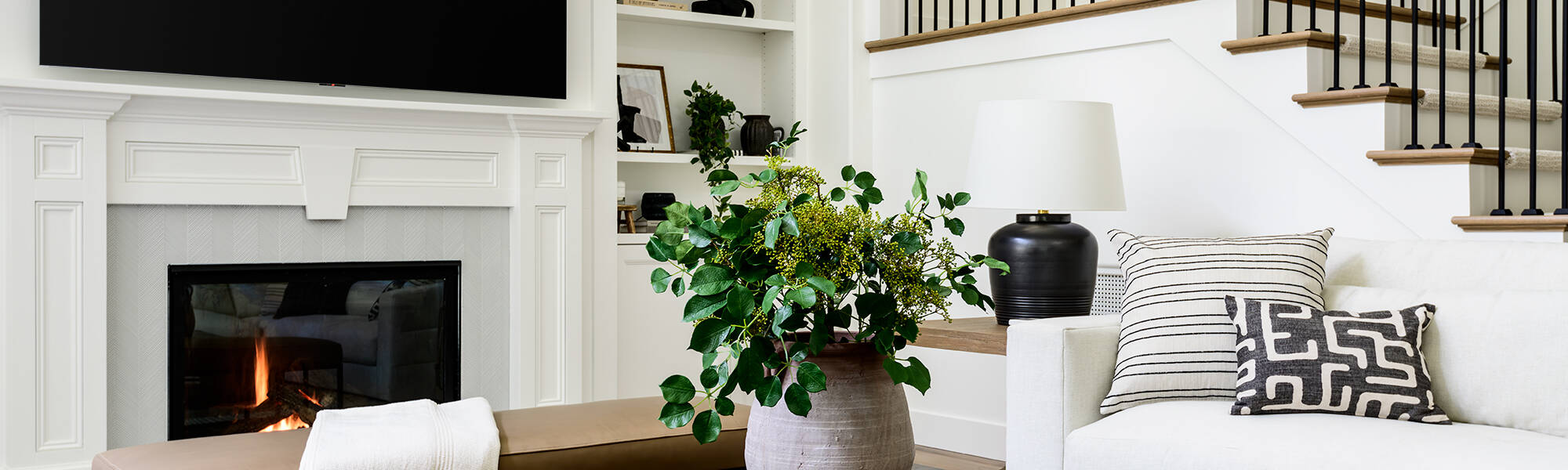 Modern living room with a fireplace, large wall-mounted TV, light-colored sofa, patterned pillows, a coffee table with decor, a potted plant, and a staircase with black railings in the background.