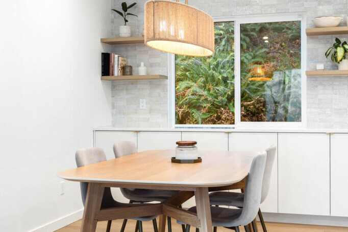 A modern dining area with a light wood table, four gray upholstered chairs, a large pendant light above, built-in white cabinets, wall shelves with decor, and a window showing greenery outside.