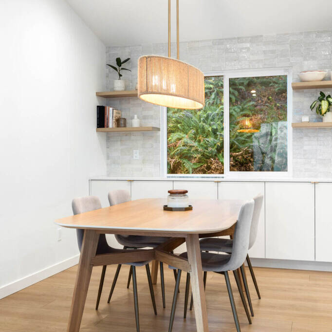 A modern dining area with a light wood table, four gray upholstered chairs, a large pendant light above, built-in white cabinets, wall shelves with decor, and a window showing greenery outside.