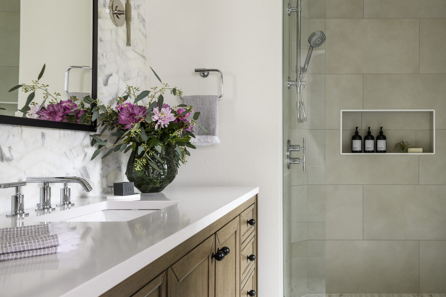 Modern bathroom with a wooden vanity, marble countertop, floral arrangement, large mirror, glass-enclosed shower, tiled walls, and shelves holding bottles and a small plant. Soft, natural lighting fills the space.