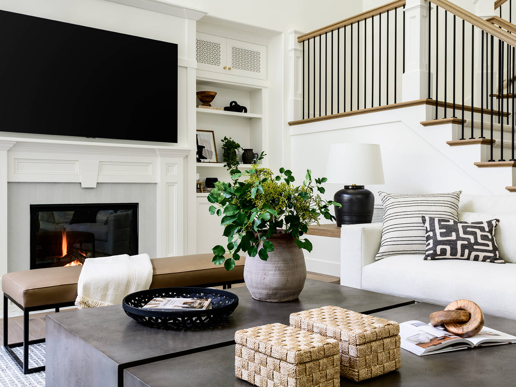 A modern living room with a white sofa, black and white pillows, a fireplace, a large TV above it, a potted plant on a coffee table, woven baskets, and a staircase with black railings in the background.