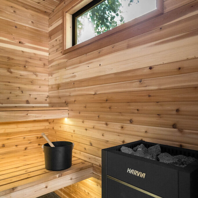 A modern wooden sauna interior with a bench, a black bucket with a ladle, and a black Harvia heater with rocks. Natural light enters through a rectangular window above.