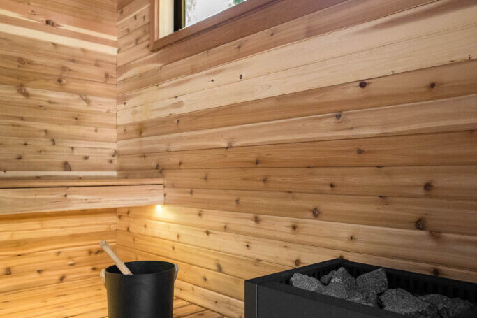 A modern wooden sauna interior features a bench with a black bucket and ladle, a Harvia heater filled with sauna rocks, wood-paneled walls, and a rectangular window letting in natural light.