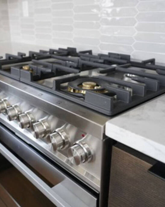 A modern stainless steel gas stove with five burners and control knobs, set in a kitchen with white hexagonal tile backsplash and marble countertops.