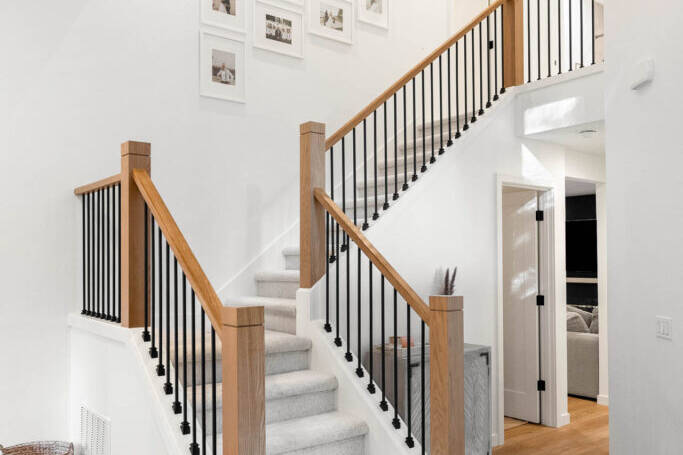 A modern staircase with light wood steps and black railings, surrounded by white walls. Framed photos hang on the wall above. A black circular chandelier hangs from the ceiling. The floor is light wood.