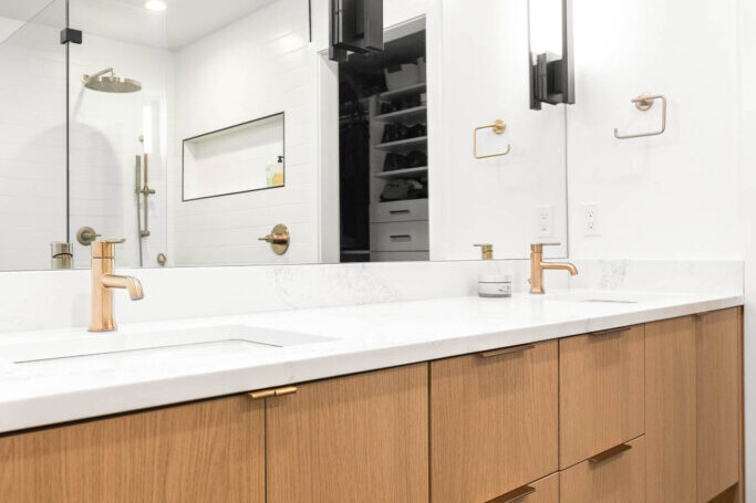 Modern bathroom with a wooden vanity, white countertop, two gold faucets, large mirror, vertical lights, and a glass shower in the background; bright and minimalistic design.