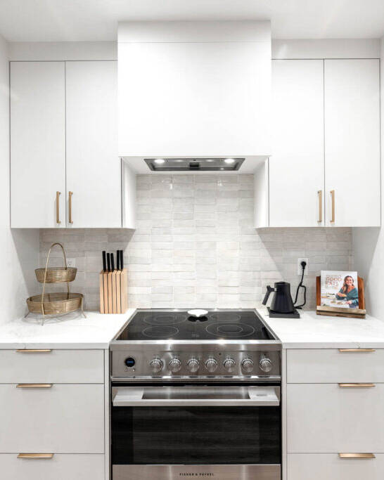 Modern kitchen with white cabinets, stainless steel oven, electric stovetop, marble backsplash, and minimal counter decor including a knife block, basket, electric kettle, and a cookbook on a stand.
