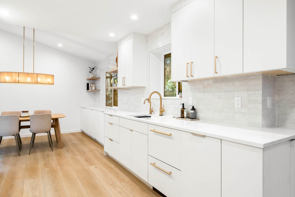 Modern kitchen with white cabinets, gold hardware, light wood flooring, and a built-in sink. A dining area with a wooden table and upholstered chairs is visible in the background under a rectangular pendant light.