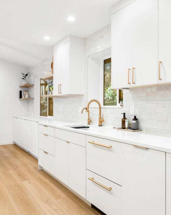 Modern kitchen with white cabinets, gold handles, a gold faucet, and light wood floors. In the background, a dining area features a wooden table, beige chairs, and a large rectangular pendant light.