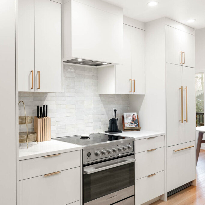 Modern kitchen with white cabinets, gold handles, stainless steel oven, knife block, kettle, cookbook, and light wood flooring. The backsplash features light gray tiles and the space is brightly lit with recessed lighting.