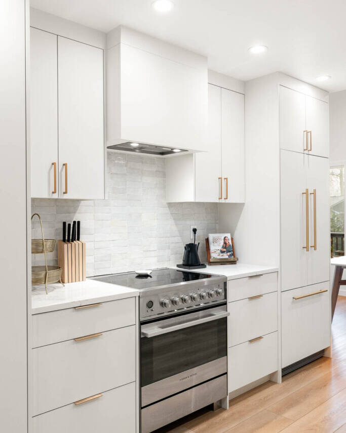 Modern kitchen with white cabinets, gold handles, stainless steel oven, knife block, kettle, cookbook, and light wood flooring. The backsplash features light gray tiles and the space is brightly lit with recessed lighting.