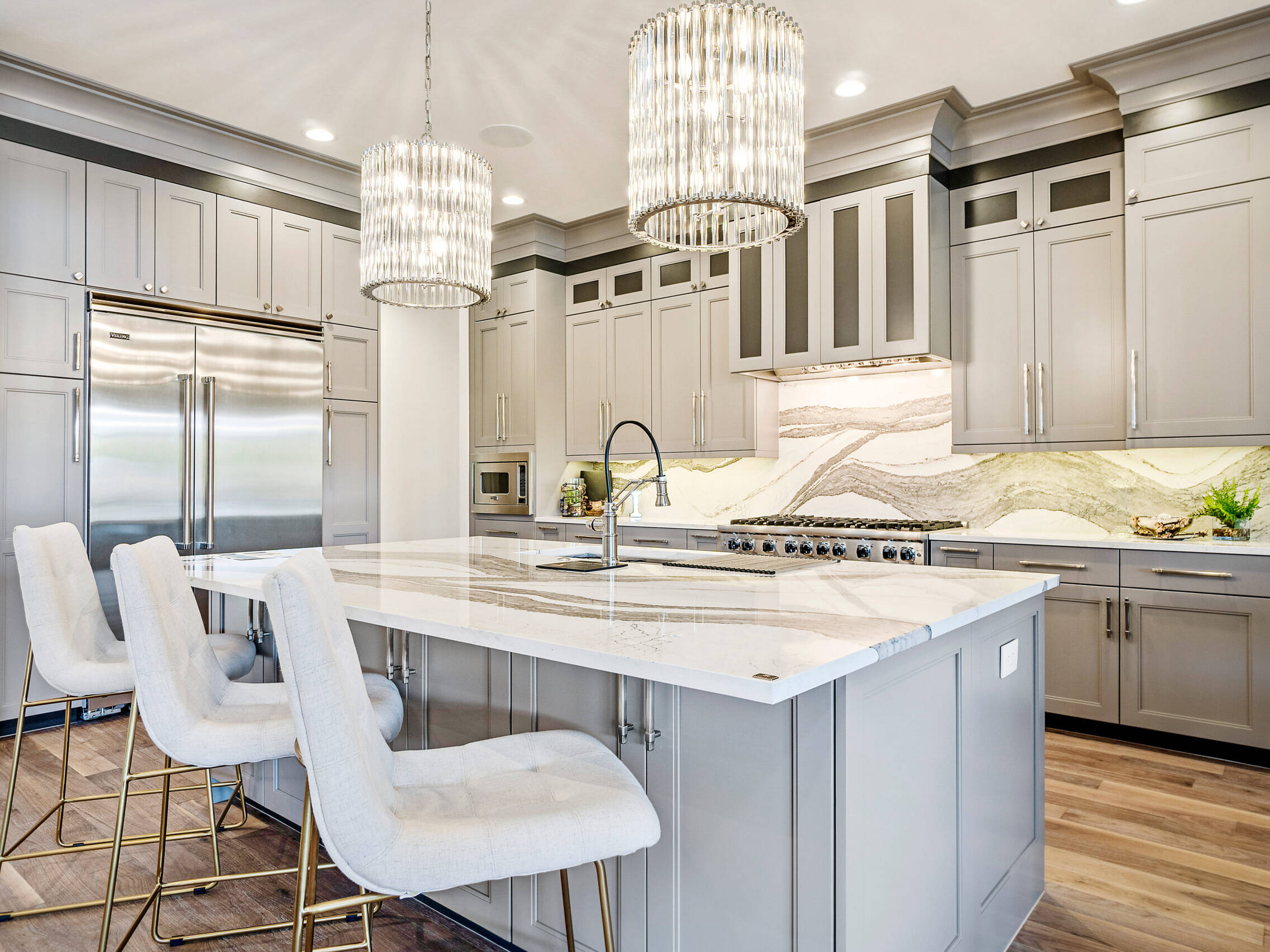 Modern kitchen with gray cabinets, marble countertops, stainless steel appliances, a large island with four upholstered chairs, pendant lights, wood flooring, and a decorative backsplash.