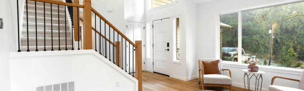 A modern, bright living room with light wood floors, white walls, a round coffee table, two armchairs, a large window, and an open staircase with wooden handrails and black balusters. A woven basket sits near the stairs.