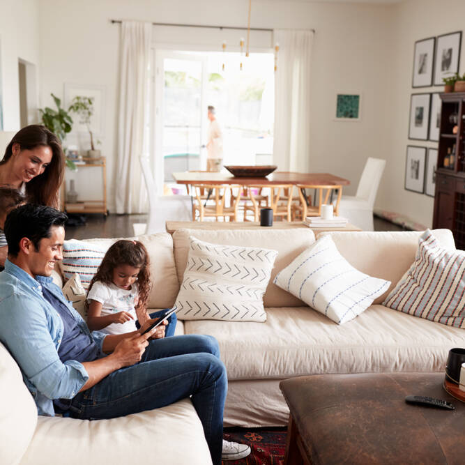A family of four relaxes in a bright living room. A man and young girl sit on a sofa using a tablet, while a woman smiles, holding a baby behind them. The room is decorated with pillows and framed art.
