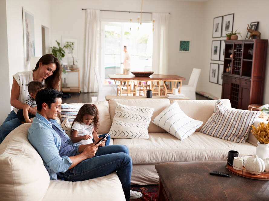 A family of four relaxes in a bright living room. A man and young girl sit on a sofa using a tablet, while a woman smiles, holding a baby behind them. The room is decorated with pillows and framed art.