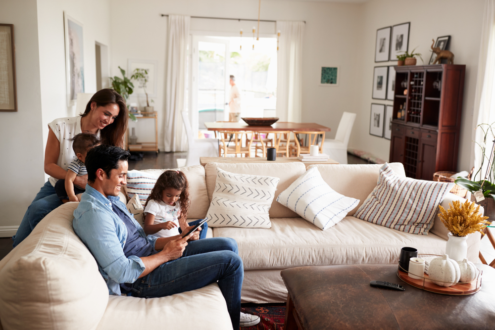 A family of four relaxes in a bright living room. A man and young girl sit on a sofa using a tablet, while a woman smiles, holding a baby behind them. The room is decorated with pillows and framed art.