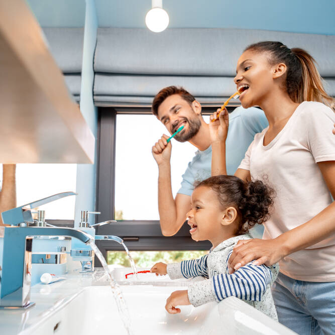 A smiling family of three, including a man, woman, and young girl, brush their teeth together at a bathroom sink. The room is bright with blue walls and large windows in the background.