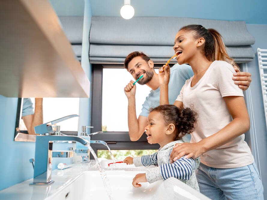 A smiling family of three, including a man, woman, and young girl, brush their teeth together at a bathroom sink. The room is bright with blue walls and large windows in the background.