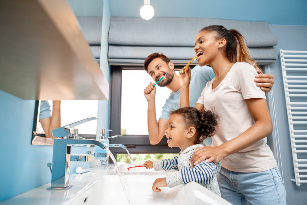 A smiling family of three, including a man, woman, and young girl, brush their teeth together at a bathroom sink. The room is bright with blue walls and large windows in the background.