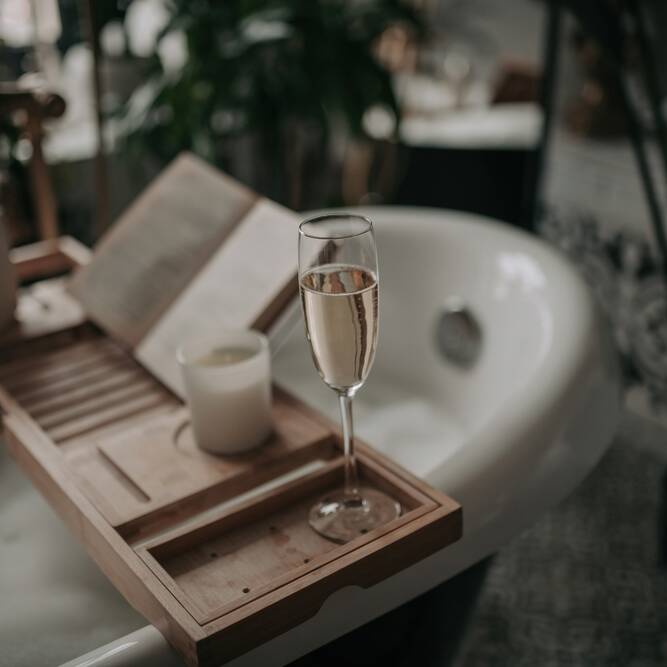 A glass of champagne, a candle, and an open book rest on a wooden bath tray across a bubble-filled bathtub, creating a relaxing and cozy atmosphere.