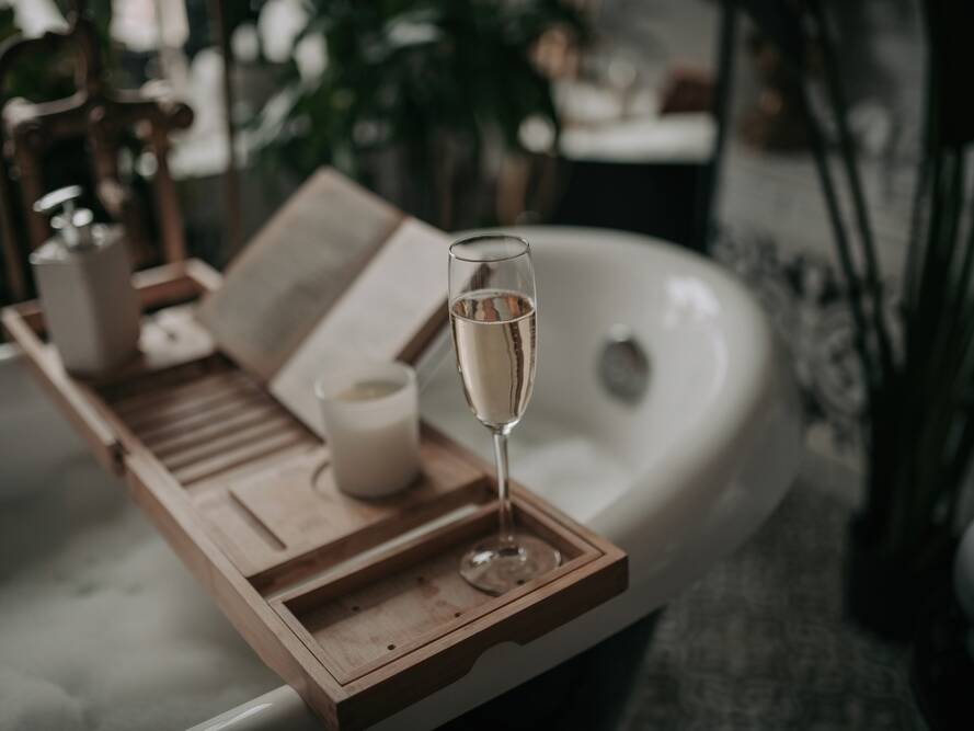 A glass of champagne, a candle, and an open book rest on a wooden bath tray across a bubble-filled bathtub, creating a relaxing and cozy atmosphere.
