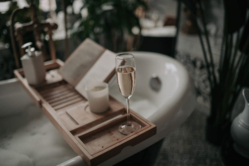 A glass of champagne, a candle, and an open book rest on a wooden bath tray across a bubble-filled bathtub, creating a relaxing and cozy atmosphere.