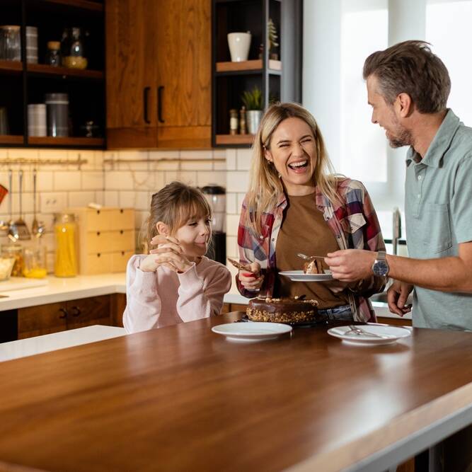 A smiling family of three stands in a modern kitchen, preparing to cut and serve a chocolate cake together, enjoying a happy moment.