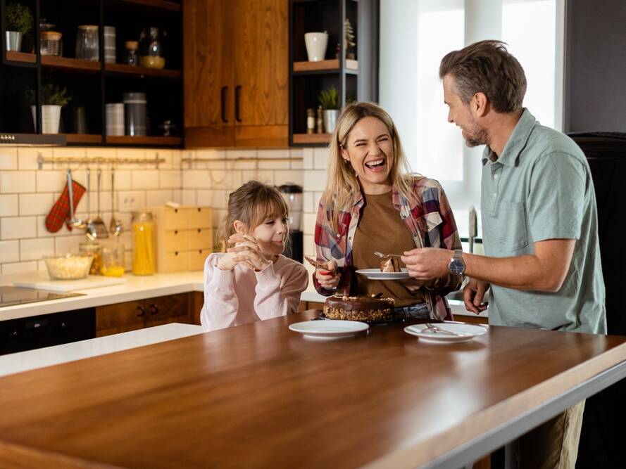 A smiling family of three stands in a modern kitchen, preparing to cut and serve a chocolate cake together, enjoying a happy moment.