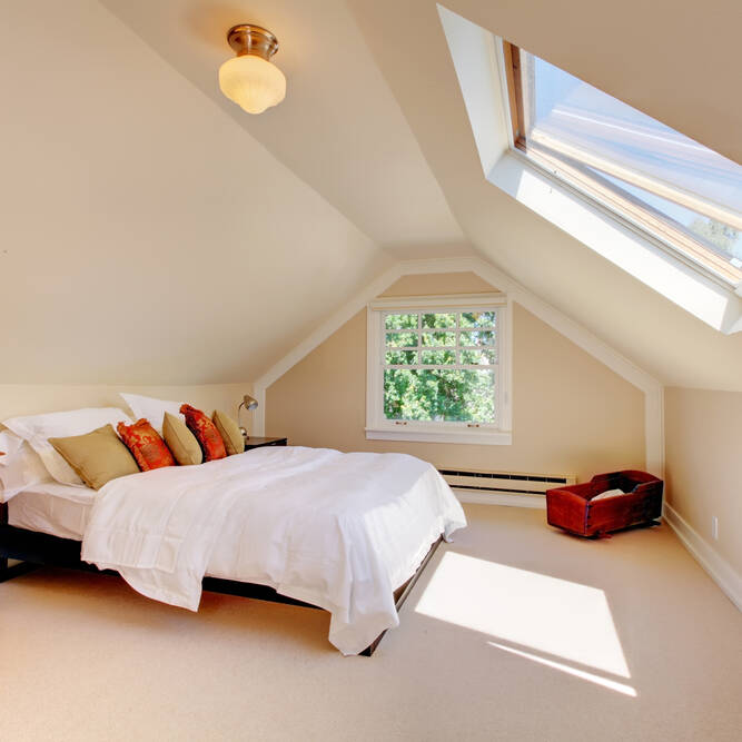 A cozy attic bedroom with a bed, white bedding, colorful pillows, a wooden chest of drawers, a skylight, and a small window letting in sunlight. A wooden chest sits near the window on a beige carpeted floor.